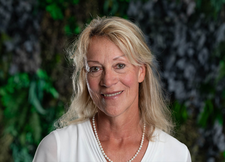 Portraitfoto von einer blonden Frau mit schulterlangen Haaren vor einer grünen Pflanzenwand.