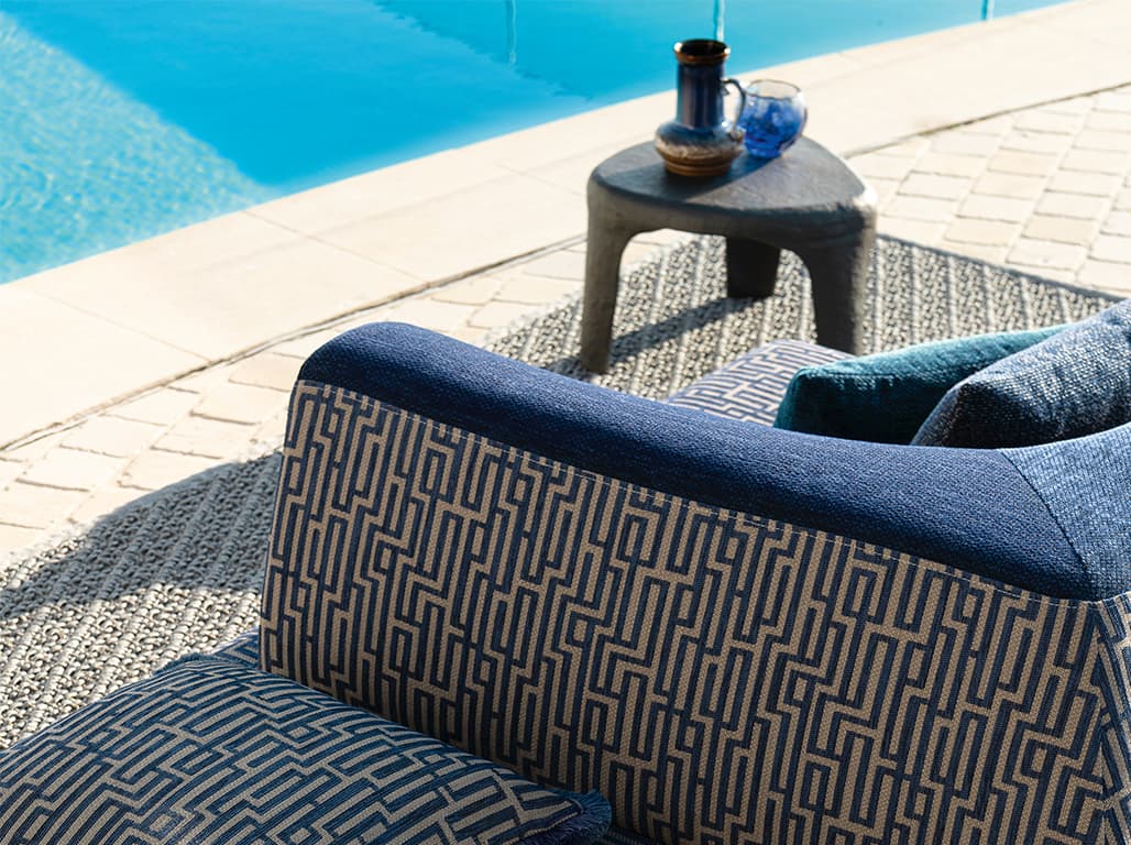 Das Bild zeigt ein blau-weiß gemustertes Sofa am Pool. Darunter liegt ein Teppich, und ein passend gefärbter Beistelltisch ergänzt die Einrichtung. Die Blau- und Weißtöne des Sofas harmonieren gut mit der Farbe des Pools.